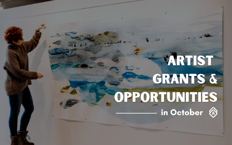 Artist Ana Žanić stands beside a large-scale abstract painting mounted on a white wall in her studio. She wears a brown knit sweater and dark pants, gesturing toward the artwork with her right hand raised. The painting features flowing organic forms in blues, whites, and earth tones, resembling a landscape or seascape with scattered dark elements throughout. The composition has a fluid, painterly quality with sweeping brushstrokes and color gradients. Text overlay reads "ARTIST GRANTS & OPPORTUNITIES in October" in white letters. The studio space has white walls and wooden ceiling beams, creating a clean, professional environment for displaying the large contemporary artwork.