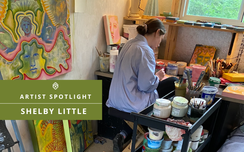 Shelby Little painting in her art studio surrounded by vibrant, myth-inspired artwork and jars of paint. Featured Artist on Artwork Archive, Shelby’s workspace is filled with colorful portraits, brushes, and in-progress canvases, reflecting her unique style rooted in memory, mythology, and dreamlike imagery.