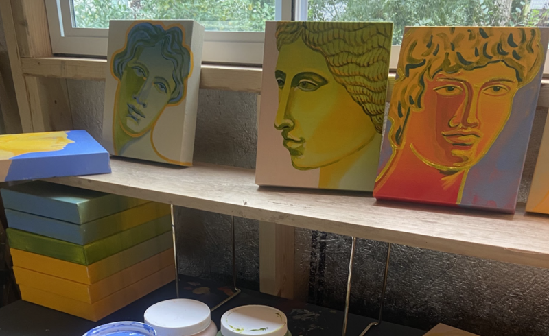 Close-up of three Greco-Roman inspired portraits by artist Shelby Little, displayed on a wooden shelf in her studio. The vibrant canvases feature bold color palettes and classical profiles, surrounded by stacked gradient-colored panels and paint supplies. Each portrait reflects a deep sense of history and is part of Little’s evolving archive of mythological and memory-driven artwork. Photo courtesy of the artist.