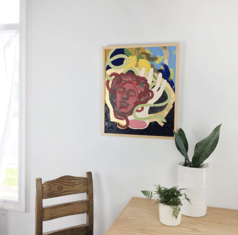 Shelby Little, Holding Onto What’s Left, 24 x 18 x 2 in. A framed painting featuring a mythic red-toned face surrounded by swirling gold, green, and blue abstract forms, hanging on a white wall above a light wood table. The artwork blends figuration with symbolism and is part of Little’s ongoing exploration of memory and myth. A rustic wooden chair and two white ceramic planters with green plants complete the serene, minimalist interior setting.